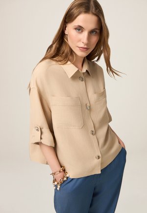 Camicia beige con bottoni, due tasche sul petto, maniche corte arrotolate e tessuto liscio. Indossata con pantaloni blu e decorata con un bracciale d'oro.