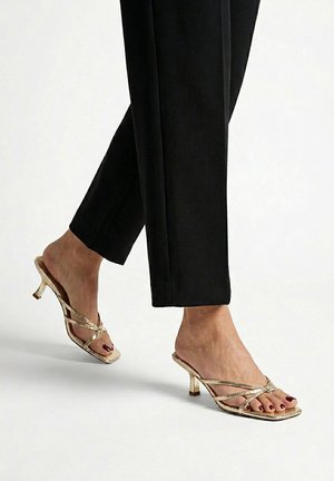 Pies con sandalias doradas de tiras y tacón alto combinadas con pantalones negros cortos sobre un fondo claro.