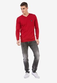 Roter V-Ausschnitt-Pullover aus Strickmaterial, lässige Passform, mit einem kleinen schwarzen Logo-Patch auf der Brust. Trägt dazu verblasste graue Jeans und weiße Sneaker.