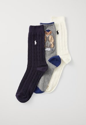Trois paires de chaussettes : en tricot torsadé violet foncé, grises avec un motif d'ours, et crème avec un design en tricot torsadé. Toutes comportent des accents de logo.