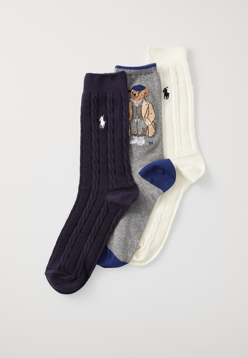 Tres pares de calcetines: uno de color púrpura oscuro con punto de cable, otro gris con un gráfico de oso y otro en crema con diseño de punto de cable. Todos tienen detalles del logo.