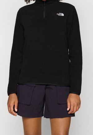 Person iført en sort kvart-lynlås North Face fleece og sorte cargoshorts, stående mod en ensfarvet lys baggrund.