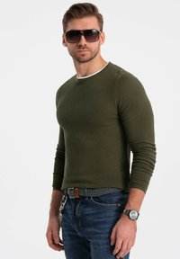 Olijfgroen gebreid T-shirt met lange mouwen en een witte halslijn, gecombineerd met blauwe jeans en een patroonriem. Model draagt een zonnebril en een horloge.