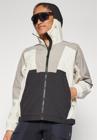 Chaqueta con bloques de color en secciones negra, beige y crema. Cuenta con un cierre de cremallera, puños elásticos y capucha. Textura de tela suave.