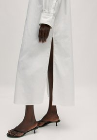 Weißes langes Hemdkleid mit einem seitlichen Schlitz, geknöpften Manschetten und lockerem Schnitt. Kombiniert mit schwarzen, hochhackigen Flip-Flops. Glatter Baumwollstoff.