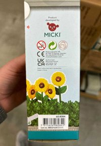 Pappschachtel vor einem hellblauen Hintergrund, dekoriert mit Sonnenblumen und grünem Laub; enthält einen Barcode sowie die CE-/UKCA-Zertifizierungszeichen.
