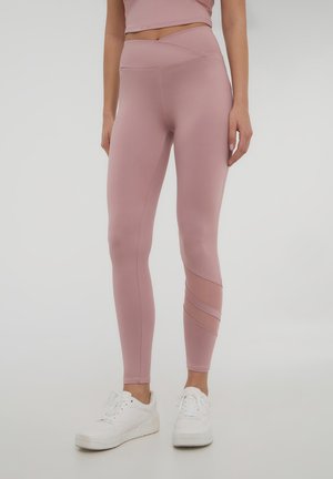 Leggings rosa con una texture liscia, dotati di una vita incrociata e di un pannello in rete con linee diagonali su una gamba. Abbinati a sneakers bianche.