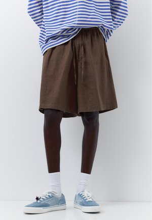 Person iført løse brune shorts, hvide ankelstrømper, lyseblå sneakers og en blå- og hvidstribet skjorte mod en ensfarvet baggrund.