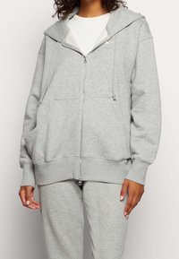 Person som bär en ljusgrå zip-up huvtröja och matchande sweatpants med en vit skjorta under, stående mot en enkel bakgrund.