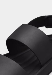 Μαύρο σανδάλι ανοιχτού δαχτύλου με δύο φαρδιές δερμάτινες λουρίδες και μαλακό πέλμα, πάνω σε απλό φωτεινό φόντο.