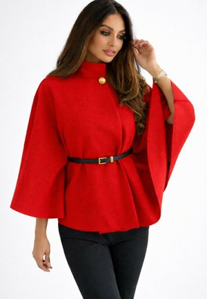 Femme portant un haut style cape rouge vif avec un bouton doré, une ceinture noire et un pantalon noir, ajustant ses cheveux d'une main.