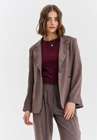 Brązowa teksturowana marynarka o lekko luźnym kroju, z wyciętymi klapami i pojedynczym zapięciem na guzik, zestawiona z bordową bluzką.