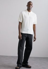 Polo shirt blanc texturé à manches courtes, associé à un jean droit noir et des sandales noires, se tenant devant un fond neutre.