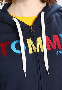 Navy zip-up hoodie featuring multicolored text "TOMMY" in red, blue, yellow, with white drawstrings and textured fabric.