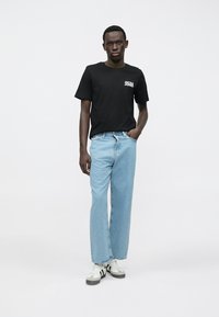 Svart T-skjorte med korte ermer og hvit logo, kombinert med vide, lyseblå jeans og hvite joggesko med svarte striper.