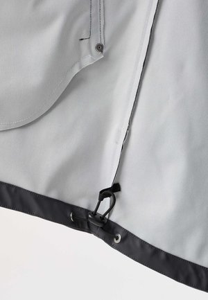 Lys gråt stof med sort kant har en sort justerbar snor og en trykknap nær kanten af materialet.