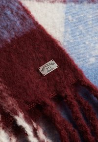 Bufanda de cuadros que presenta una mezcla de colores burdeos, azul y blanco. Fabricada en un tejido suave y peludo, con bordes flecados y una etiqueta de marca metálica.