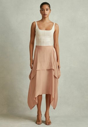 Frau mit zurückgebundenem Haar, die ein cremefarbenes, figurbetontes ärmelloses Oberteil und einen mehrlagigen High-Low-Rock in Blush-Rosa mit naturfarbenen offenen Absatzschuhen trägt.