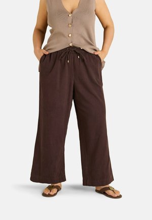 Persona que lleva pantalones anchos y sueltos de color marrón oscuro con cintura ajustable, top sin mangas marrón claro con botones y sandalias marrones, manos en los bolsillos.