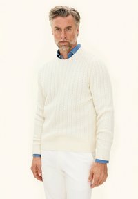 Biały sweter w sploty z okrągłym dekoltem, noszony na jasnoniebieskiej koszuli z kołnierzykiem, w zestawie z białymi spodniami. Widoczny teksturowany wzór splotu.