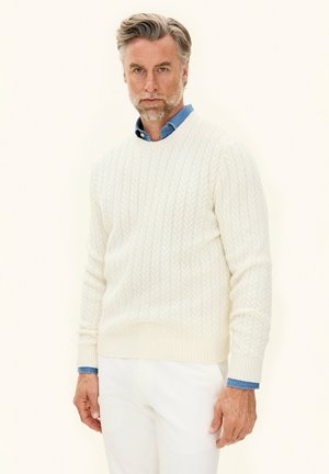 Maglione bianco a maglia cable-knit con scollatura rotonda, indossato sopra una camicia colletto azzurra, abbinato a pantaloni bianchi. Il motivo a maglia strutturato è visibile.