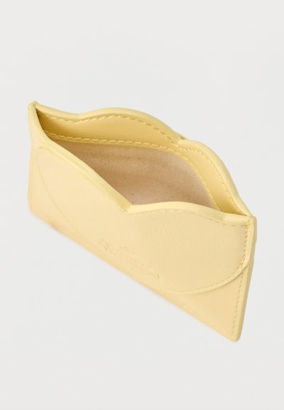 Κίτρινο δερμάτινο θήκη καρτών με κυματιστό πάνω άκρο, ραμμένες λεπτομέρειες και λεία επιφάνεια. Το εσωτερικό διαθέτει ελαφριά υφασμάτινη επένδυση.