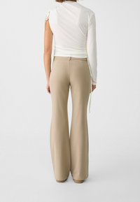 Vrouw in beige flare-broek en een witte asymmetrische top met lange mouwen, staande met de rug naar de camera tegen een eenvoudige achtergrond.