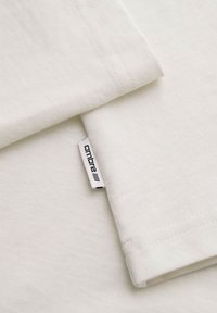 Tissu en coton blanc avec des coutures visibles et une petite étiquette blanche portant la mention « ombre » attachée au bord.