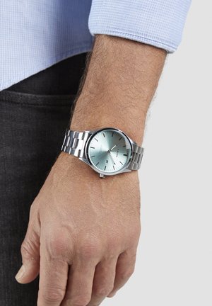 Silberne Metall-Armbanduhr mit rundem, hellblauem Zifferblatt und silbernen Stundenmarkierungen, auf einem silbernen Gliederarmband getragen, am Handgelenk mit heller Haut.