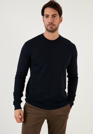 Navy blue knitted sweater with a textured pattern, crew neckline, and ribbed cuffs and hem. Worn with brown trousers.