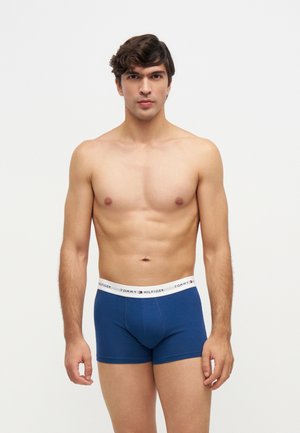 Jeune homme debout, face à l’avant, portant un boxer bleu Tommy Hilfiger avec une ceinture blanche sur fond uni.