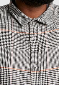 Camisa de cuadros en blanco y negro con acentos de cuadro naranja, con un cuello estructurado y cierre frontal de botones, confeccionada en tela tejida.
