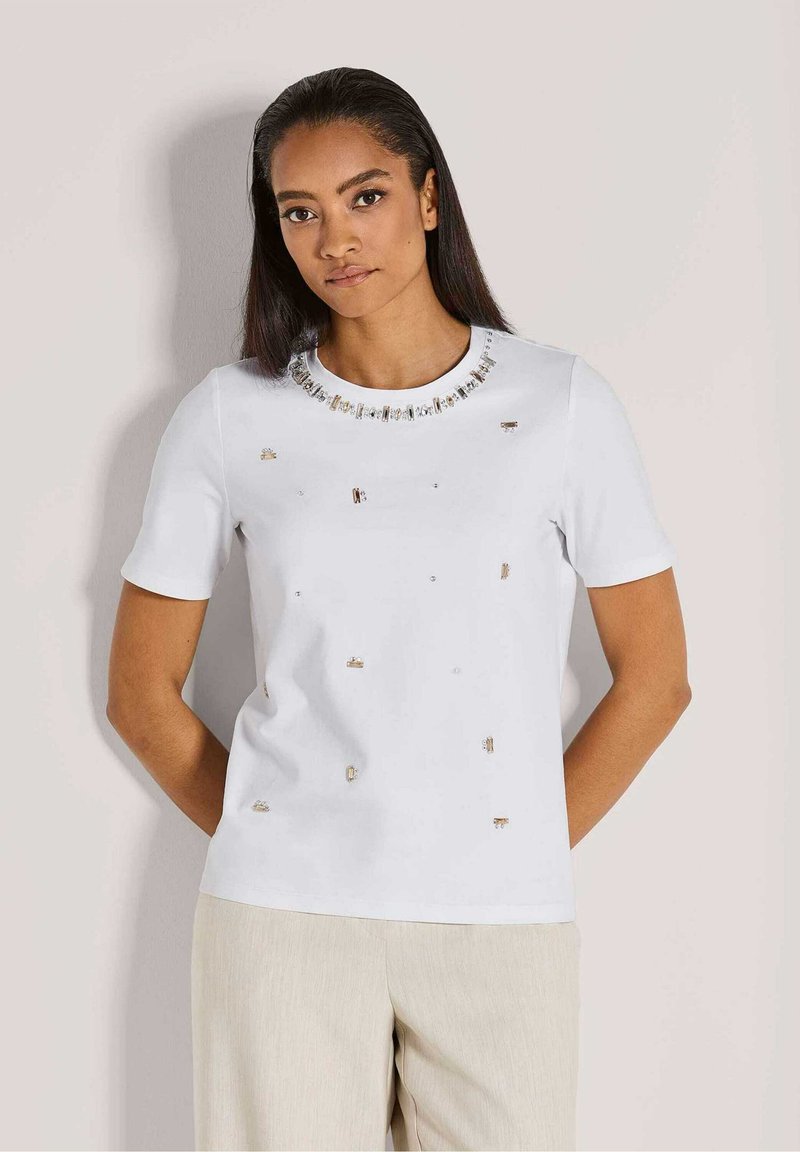 Witte kortekraagtop met ronde hals. Heeft decoratieve versieringen en een gladde textuur. Gepaarde met lichte broeken.