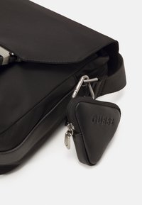 Borsa a tracolla nera in nylon con chiusura a patta, dotata di una tasca triangolare con zip e dettagli in metallo argentato. Dettaglio della superficie testurizzata presente.