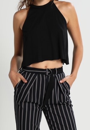 Kvinde iført sort halter crop top og højtaljede sorte bukser med hvide lodrette striber samt sort bælte med rund spænde.