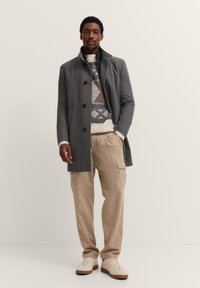 Grau gestrickter Wollmantel mit hohem Kragen, gemustertem creme- und blauen Pullover, beigen Cargo-Hosen und hellen Sneakers. Glatte Textur insgesamt.