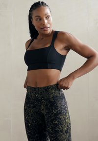 Sujetador deportivo negro con tirantes anchos y costuras sutiles, combinado con leggings de cintura alta que presentan un patrón abstracto en verde y negro.