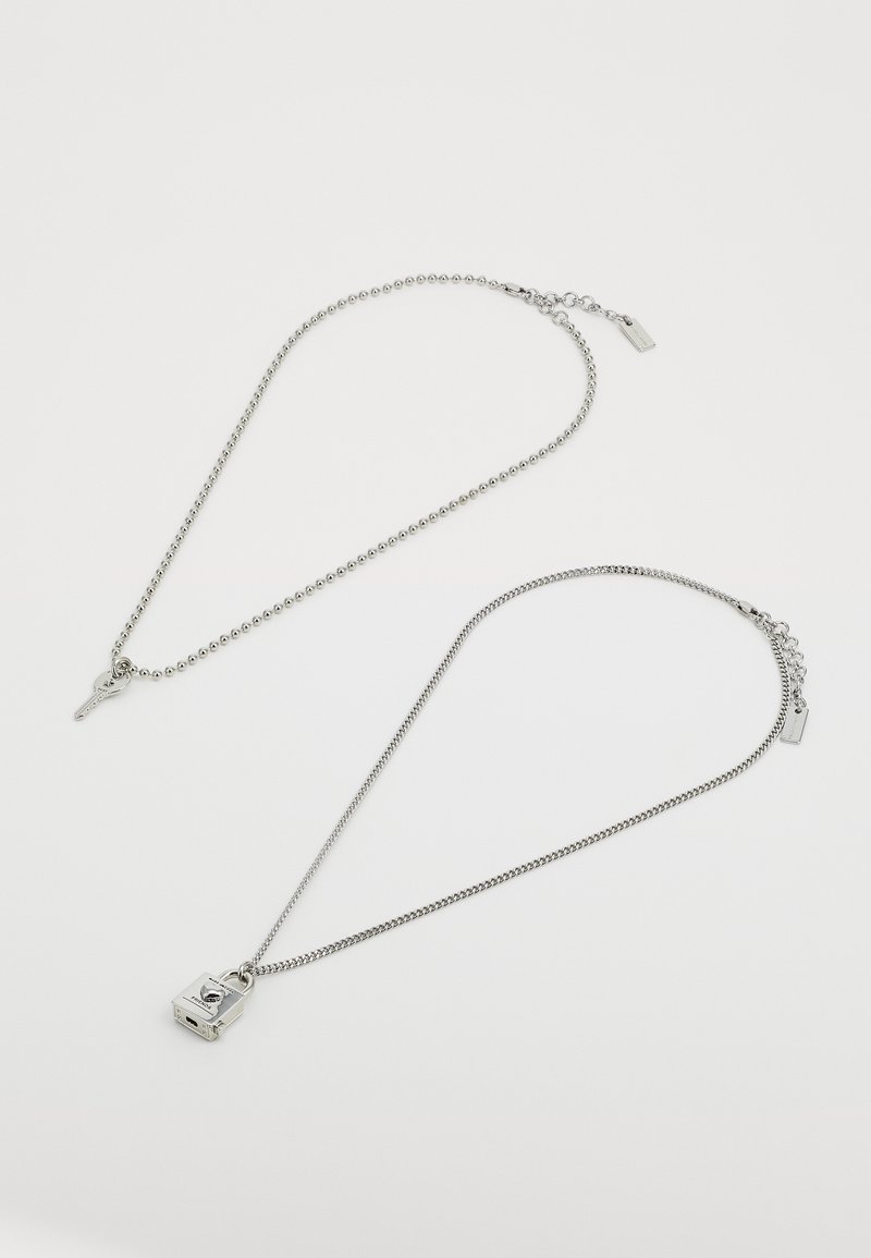 Prikazane su dvije srebrne narukvice: jedna s kuglastim lančićem i privjeskom u obliku ključa, a druga s privjeskom s kutijom i glatkim lančićem.