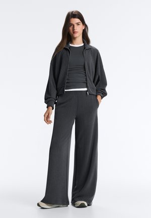 Tânără în picioare, purtând o jachetă gri închis cu fermoar, pantaloni largi asortați, un tricou negru peste un top alb și adidași albi.