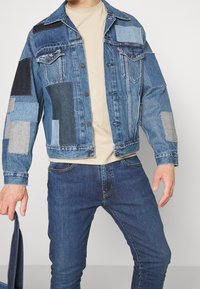 Džínová bunda s různými kousky nášivek v odstínech modré a šedé, s kovovými knoflíky a klasickým límcem.