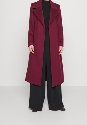 Personne portant un long manteau bordeaux par-dessus un haut noir et un pantalon noir à jambes larges, se tenant devant un fond clair uni.