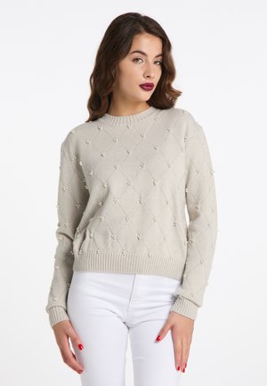 Donna che indossa un maglione beige chiaro con piccoli dettagli di perle e pantaloni bianchi, che posa con una mano appoggiata sulla coscia.