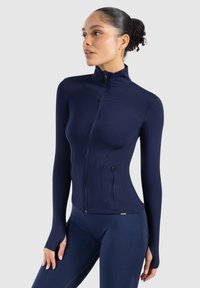 Chaqueta atlética de color marino con cuello alto, cierre de cremallera completa, diseño ajustado y agujeros para los pulgares en las mangas. Tejido suave y elástico con bolsillo lateral.