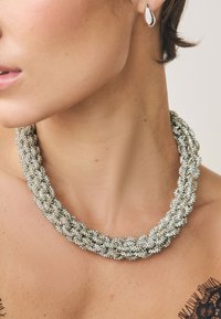 Silver beaded necklace with braided design, featuring multiple strands for texture and a smooth finish. Small decorative hardware accents visible.