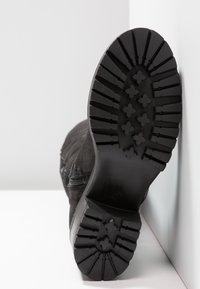 Bota de ante negro con suela gruesa de goma que muestra surcos profundos y agarres en forma de cruz, apoyada contra una superficie blanca.