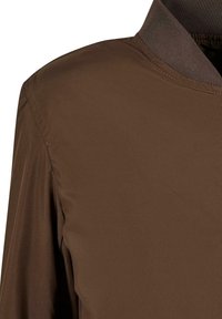 Giacca brown leggera con una texture liscia e un design semplice. Il colletto è scuro e i dettagli di cucitura sono visibili lungo le cuciture.