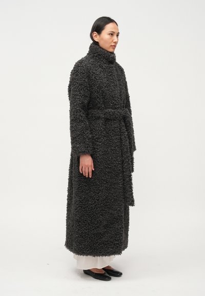 Dugi tamnosivi kaput od teksturirane, pahuljaste tkanine s visokim ovratnikom. Ima pojas za vezanje i bočne džepove, nošen preko svijetle odjeće.