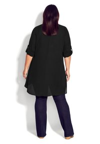 Tunique noire à manches retroussables, ourlet asymétrique et empiècements en tissu transparent à l'arrière, associée à un jean évasé bleu foncé et des chaussures couleur beige.