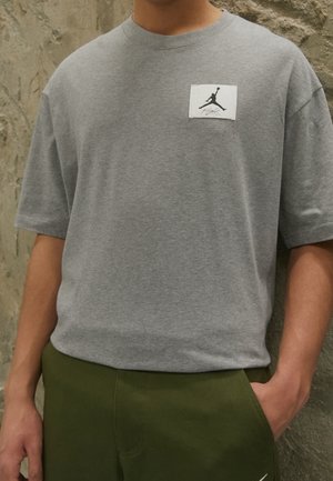 Osoba nosząca szarą koszulkę z małą czarną naszywką z logo Jumpman i oliwkowozielone spodnie, z jedną ręką w kieszeni.