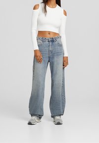 Top blanco de manga larga recortado con hombros descubiertos, combinado con jeans de pierna ancha azul claro y zapatillas grises. Diseño simple y textura de tela.